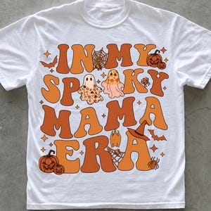 Könnte beinhalten: Ein weißes Kurzarm-T-Shirt mit einem großen Grafikdruck in Orange- und Brauntönen. Der Text lautet IN MY SPOOKY MAMA ERA, umgeben von Illustrationen von Geistern, Kürbissen, Spinnen, Spinnennetzen, Fledermäusen und einem Hexenhut.