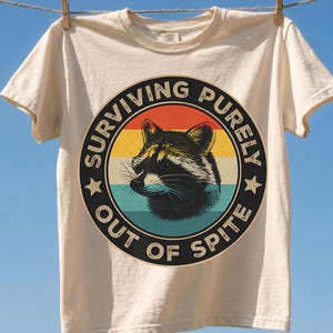 Könnte beinhalten: Ein cremefarbenes Kurzarm-T-Shirt mit einer kreisförmigen Grafik, die einen Waschbären zeigt. Das Design enthält horizontale Streifen in Orange, Gelb und Türkis sowie den in Schwarz gedruckten Text "SURVIVING PURELY OUT OF SPITE".