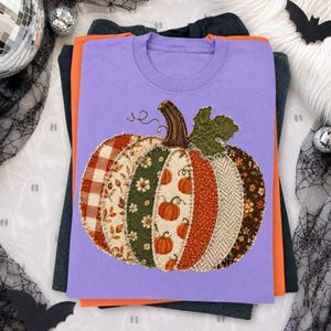 Könnte beinhalten: Ein hellviolettes T-Shirt mit einer großen Kürbisgrafik, die aus verschiedenen stoffartigen Flicken besteht. Das Kürbisdesign umfasst Segmente mit rotem Karomuster, Blumen- und Herbstmotiven, gekrönt von einem braunen Stiel und einem grünen Blatt.
