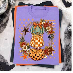 Könnte beinhalten: Ein lavendelfarbenes T-Shirt mit einer Grafik aus drei gestapelten Kürbissen, verziert mit orange-weißem Karomuster, türkisfarbenem Paisley-Muster und orangefarbenen Punkten, umgeben von einem Kranz aus Herbstblumen und orangefarbenen Sternen.