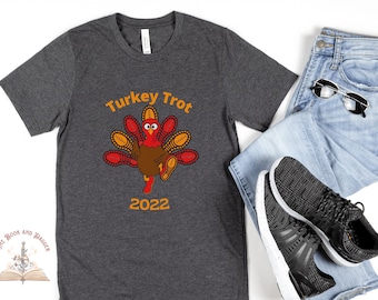 May include: Dark gray t-shirt with a cartoon turkey design. The turkey is running and the text "Turkey Trot 2022" is printed in orange. The shirt is paired with blue jeans, sunglasses, and black sneakers.