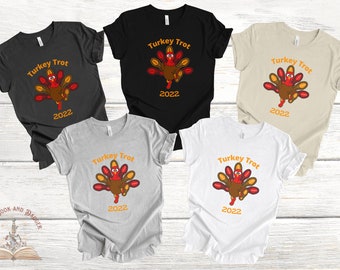 May include: Five t-shirts in various colors: black, white, grey, tan, and dark grey. Each shirt features a cartoon turkey with the text "Turkey Trot 2022" in orange. The turkey has red, brown, and yellow feathers.