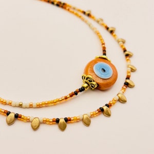 Peut inclure: Un collier à plusieurs niveaux avec une perle centrale en forme d'œil orange-brun avec un iris bleu. Le collier est composé de plusieurs brins de petites perles colorées dans les tons orange, noir et blanc, avec des breloques dorées en forme de feuille.