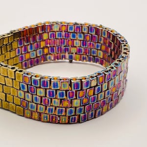 May include: A wide bracelet with a rectangular design. The bracelet is made of small, square, iridescent beads that shift in color from blue to purple to gold. The clasp and end pieces are a brassy gold color.