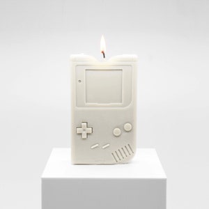 A lit candle that is modeled to look like an original Nintendo Gameboy from 1989, which was Nintendo's second ever handheld game console. The candle is displayed on top of a white museum pedestal.