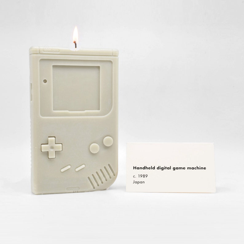 A lit candle that is modeled after the original Nintendo Gameboy. It is sitting next to a museum placard that says, "Handheld digital game machine. c. 1989. Japan."