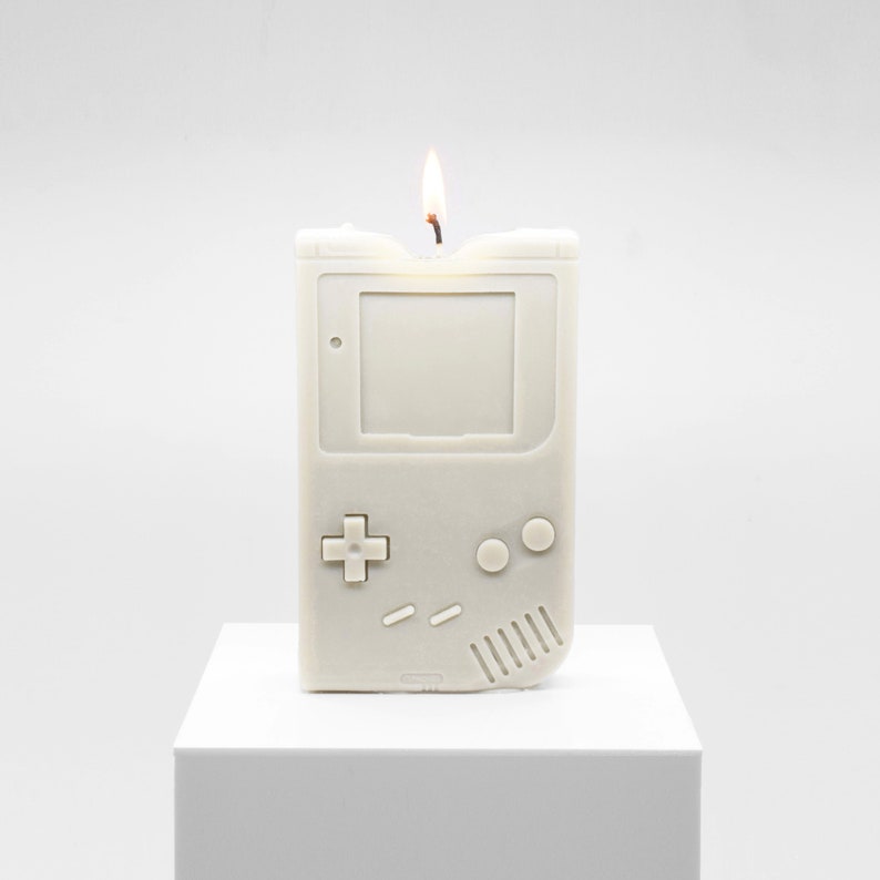 A lit candle that is modeled to look like an original Nintendo Gameboy from 1989, which was Nintendo's second ever handheld game console. The candle is displayed on top of a white museum pedestal.