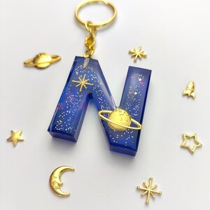 Könnte beinhalten: Ein blauer Harz-Schlüsselanhänger in Form des Buchstabens N mit goldenem Glitzer und einem goldenen Planeten mit einem Ring darum. Der Schlüsselanhänger hat auch goldene Sterne und eine goldene Rakete.