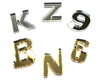 May include: A set of acrylic letters in silver and gold. The letters are K, Z, 9, B, N, and E. The letters are all uppercase and are in a simple, modern font.