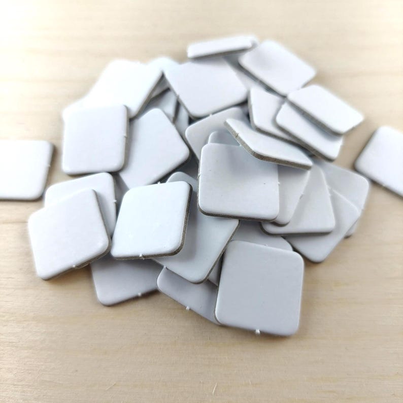 Puede incluir: Un mont&oacute;n de peque&ntilde;as almohadillas adhesivas blancas cuadradas con esquinas redondeadas. Las almohadillas tienen una superficie lisa y mate y un fino respaldo marr&oacute;n. Est&aacute;n esparcidas sobre una superficie de madera clara.