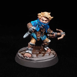 May include: Miniature figurine of a fantasy character, a dwarf or gnome, with blonde hair and a blue tunic. The figure is holding a crossbow and a dagger, standing on a cobblestone base. The figure is painted with detailed shading.