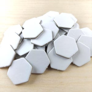 Puede incluir: Un mont&oacute;n de baldosas hexagonales blancas con un borde marr&oacute;n claro. Las baldosas est&aacute;n dispuestas sobre una superficie de madera clara. Las baldosas parecen ser de cer&aacute;mica o un material similar.