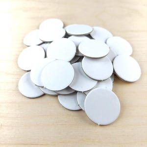 Puede incluir: Un mont&oacute;n de peque&ntilde;os c&iacute;rculos de cart&oacute;n blancos. Los c&iacute;rculos tienen una superficie blanca y un borde marr&oacute;n claro. Los objetos est&aacute;n dispersos sobre una superficie de madera clara.