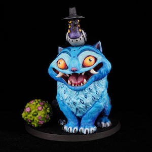 May include: A hand-painted figurine of a bright blue cat with large orange eyes, sharp teeth, and a wide grin. A small purple bird wearing a black top hat sits on the cat's head. A textured green sphere with small pink flowers sits to the left.