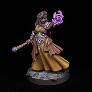 Puede incluir: Figura de fantasía en miniatura de una mujer con un vestido amarillo y marrón fluido, sosteniendo un bastón con un orbe púrpura y energía púrpura brillante en su mano. La figura tiene cabello castaño y está sobre una base de piedra.