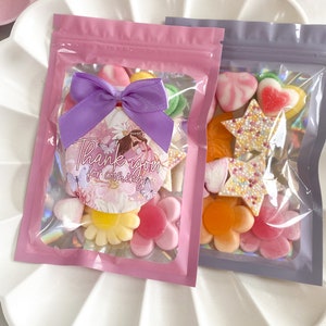 May include: Two clear plastic bags filled with colourful sweets. The pink bag has a purple bow and a round sticker that says "Thank you for coming". The purple bag has a variety of sweets, including hearts, stars, and flowers.