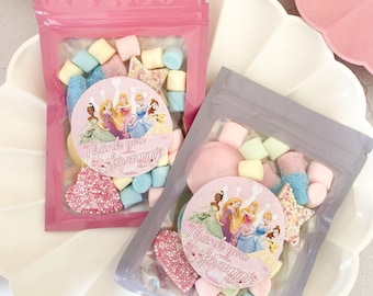 Pode incluir: Duas sacolas de plástico transparentes cheias de doces e marshmallows. As sacolas são decoradas com adesivos de princesas e o texto "Obrigado por vir".