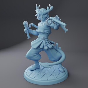 May include: A light blue figurine of a character with a horned headdress and a flowing outfit. The figure is holding two cylindrical objects and has a tail. The figurine is standing on a wooden platform.