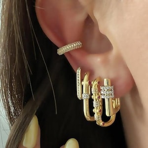 May include: A set of gold-coloured earrings and an ear cuff. The ear cuff is covered in small, clear stones. The earrings include a small hoop with clear stones, a hoop with clear and red stones, and a larger hoop with clear stones.