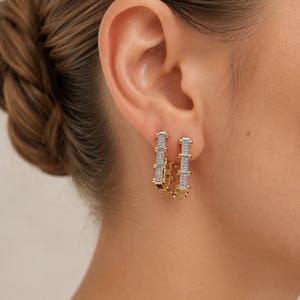 Könnte beinhalten: Ein Paar goldene Creolen mit rechteckigem Gliederketten-Design. Jeder Ohrring ist mit einer Reihe kleiner, klarer, funkelnder Steine besetzt, die in die Vorderseite eingelassen sind und für eine strukturierte, geometrische Optik sorgen.