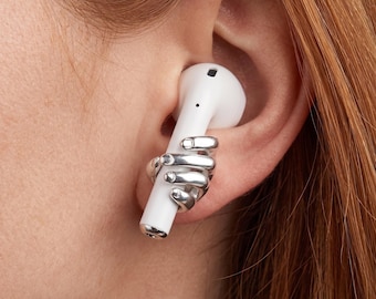May include: A white wireless earbud held by a silver hand-shaped ear cuff. The ear cuff is detailed with individual fingers and is worn on the ear. The earbud is inserted into the ear.