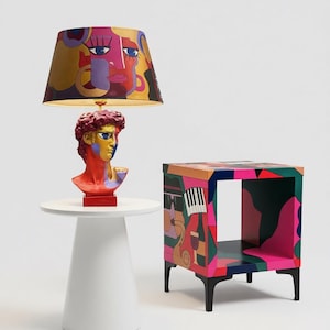 Puede incluir: Una lámpara de mesa con una pantalla de patrones abstractos coloridos y una base con forma de busto de David, pintada en rojo, dorado y azul. A su lado hay una mesa auxiliar cuadrada con un estampado gráfico multicolor y patas negras.