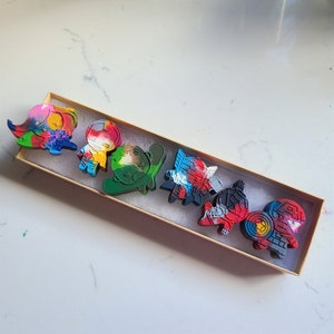 Puede incluir: Un conjunto de siete piezas de arte de crayón de colores en una caja de madera. Las piezas de arte tienen forma de superhéroes y presentan una variedad de colores y diseños.
