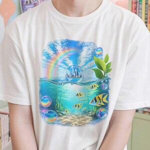 May include: A white short-sleeved t-shirt featuring a graphic print of an underwater scene with yellow and black striped fish, bubbles, a rainbow, and a small city inside a glass sphere floating above the water surface.