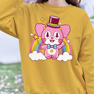 May include: A mustard yellow sweatshirt with a cartoon illustration of a pink bear wearing a top hat and bow tie. The bear is holding a magic wand and standing on a rainbow over clouds.