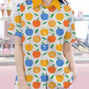 May include: A white short-sleeved button-down shirt with a yellow collar and a pattern of blue, orange, and yellow apples with faces.