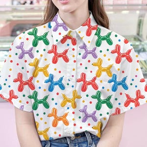 May include: A white button-down shirt with a colorful balloon animal print. The shirt has short sleeves and a button-up front. The balloon animals are in the shape of dogs and are in various colors, including red, green, blue, purple, and yellow.