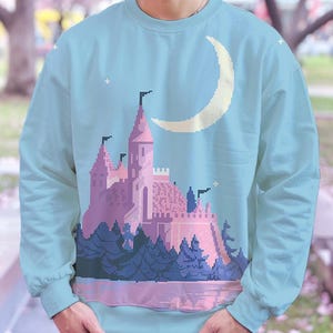 May include: A light blue sweatshirt with a pixelated design of a pink castle, trees, and a crescent moon. The design is in a retro video game style.