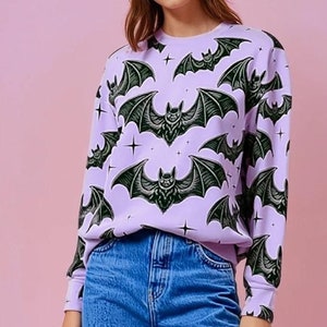May include: A light purple sweatshirt featuring a repeating pattern of stylized black bats with spread wings and star accents. The long-sleeved top is paired with blue jeans, creating a casual, everyday look. The design is suitable for Halloween or gothic-themed apparel.