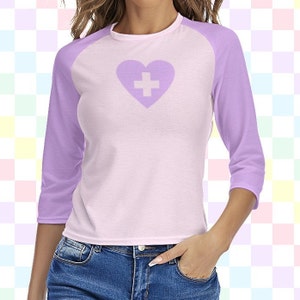 May include: A light pink and purple short-sleeved shirt with a white heart with a white plus sign in the center. The shirt has a baseball style design with a purple sleeve.