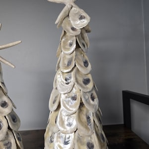 May include: A conical decorative sculpture constructed from overlapping off-white oyster shells, arranged in a tiered pattern to resemble a tree. The structure is topped with a single, light-colored starfish, creating a coastal-themed ornament.