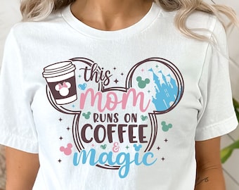 Może przedstawiać: Biała koszulka z brązowym wzorem ucha Myszki Miki. Wzór zawiera kubek kawy, tekst "This Mom Runs on Coffee & Magic" i niebieski zamek.