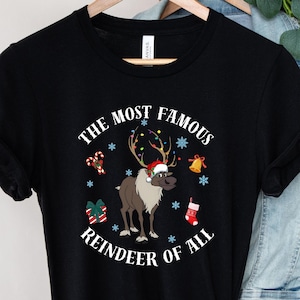 Pu&ograve; includere: Maglietta nera con una grafica bianca di una renna che indossa un cappello di Babbo Natale e circondata da decorazioni natalizie. Il testo "The Most Famous Reindeer of All" &egrave; stampato sulla maglietta.