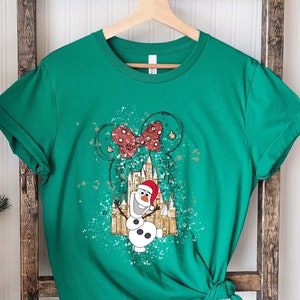 May include: Green t-shirt with a white and gold graphic of Olaf the snowman from Frozen, wearing a Santa hat, in front of a castle with a red bow on top. The graphic has a splatter paint effect.