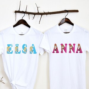May include: Two white t-shirts with the names "Elsa" and "Anna" printed on them in colorful letters. Each letter is decorated with a cartoon image of the character from the movie Frozen.
