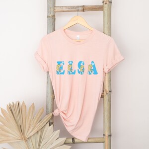 May include: A pink t-shirt with the word "ELSA" spelled out in blue letters. Each letter is decorated with a cartoon image of Elsa from the movie Frozen.