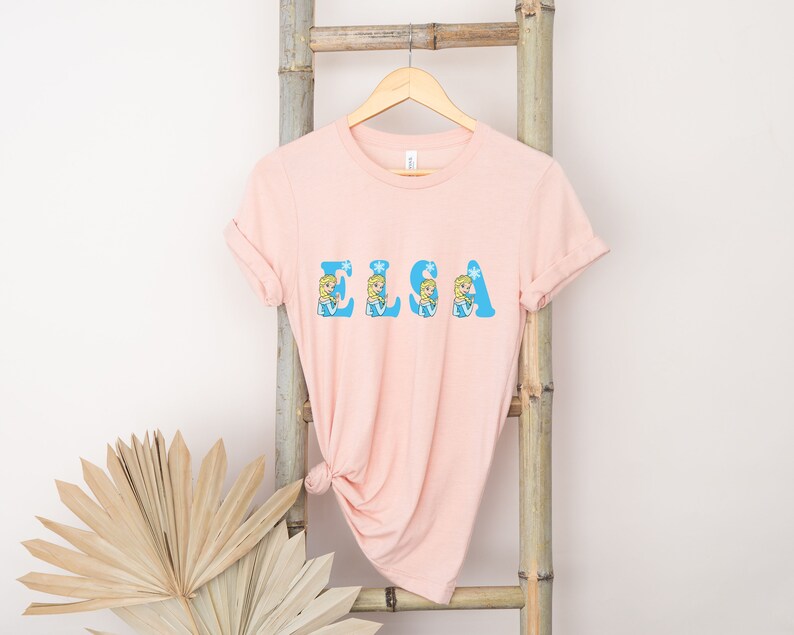 May include: A pink t-shirt with the word "ELSA" spelled out in blue letters. Each letter is decorated with a cartoon image of Elsa from the movie Frozen.