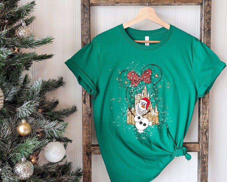 May include: Green t-shirt with a white and gold graphic of Olaf the snowman from Frozen, wearing a Santa hat, in front of a castle with a red bow on top. The graphic has a splatter paint effect.