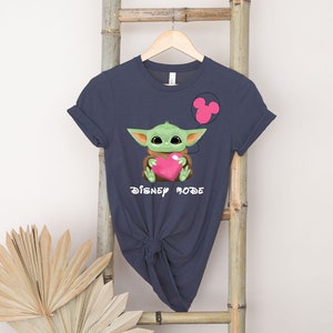 May include: A dark blue t-shirt featuring a green baby Yoda character holding a pink heart. The shirt has the text "Disney Mode" printed on it.