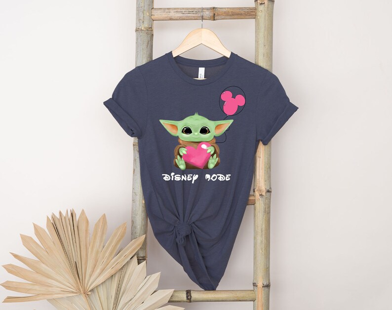 May include: A dark blue t-shirt featuring a green baby Yoda character holding a pink heart. The shirt has the text "Disney Mode" printed on it.