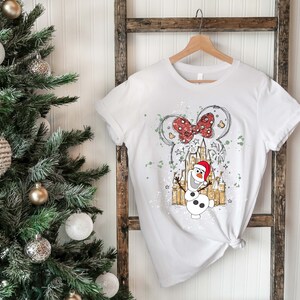 May include: White t-shirt with a graphic of Olaf the snowman from Frozen wearing a Santa hat in front of a sketch of Cinderella's castle with a red bow on top. The graphic is surrounded by white paint splatters.