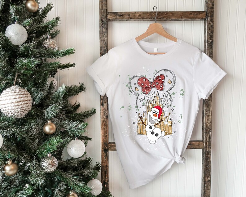 May include: White t-shirt with a graphic of Olaf the snowman from Frozen wearing a Santa hat in front of a sketch of Cinderella's castle with a red bow on top. The graphic is surrounded by white paint splatters.