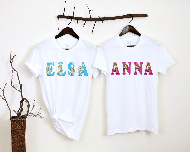 May include: Two white t-shirts with the names "Elsa" and "Anna" printed on them in colorful letters. Each letter is decorated with a cartoon image of the character from the movie Frozen.