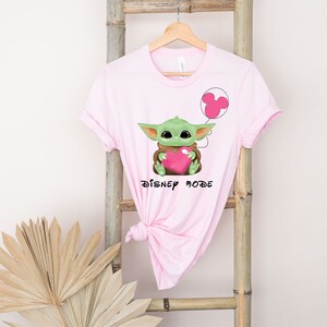 May include: Pink t-shirt featuring a green Baby Yoda character holding a pink heart and a pink Mickey Mouse balloon. The text "Disney Mode" is printed below the character.