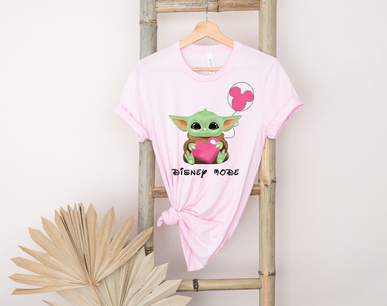 May include: Pink t-shirt featuring a green Baby Yoda character holding a pink heart and a pink Mickey Mouse balloon. The text "Disney Mode" is printed below the character.