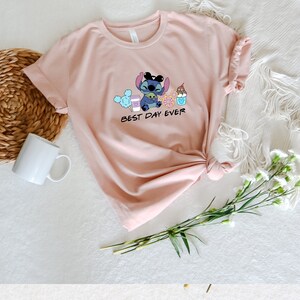 May include: A pink t-shirt with a blue cartoon character holding a cup of coffee, a donut, a Mickey Mouse ice cream cone, and a cotton candy. The text "BEST DAY EVER" is printed below the character.